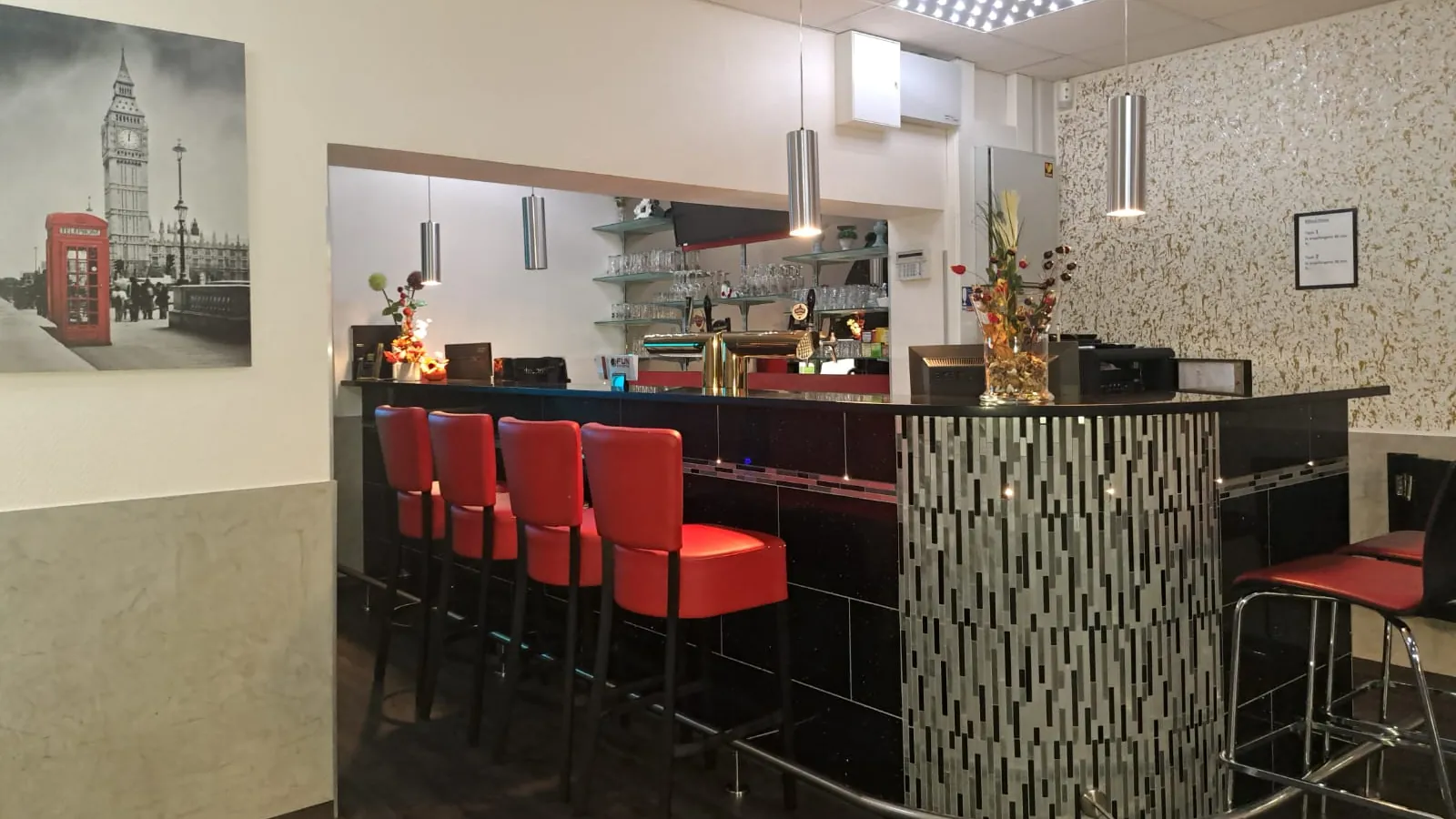 Bartheke der Sportsbar Wesel mit schwarzer Front, glitzernder Mosaiksäule, vier roten Barhockern und Pendelleuchten, dahinter Gläserregal und Zapfbereich