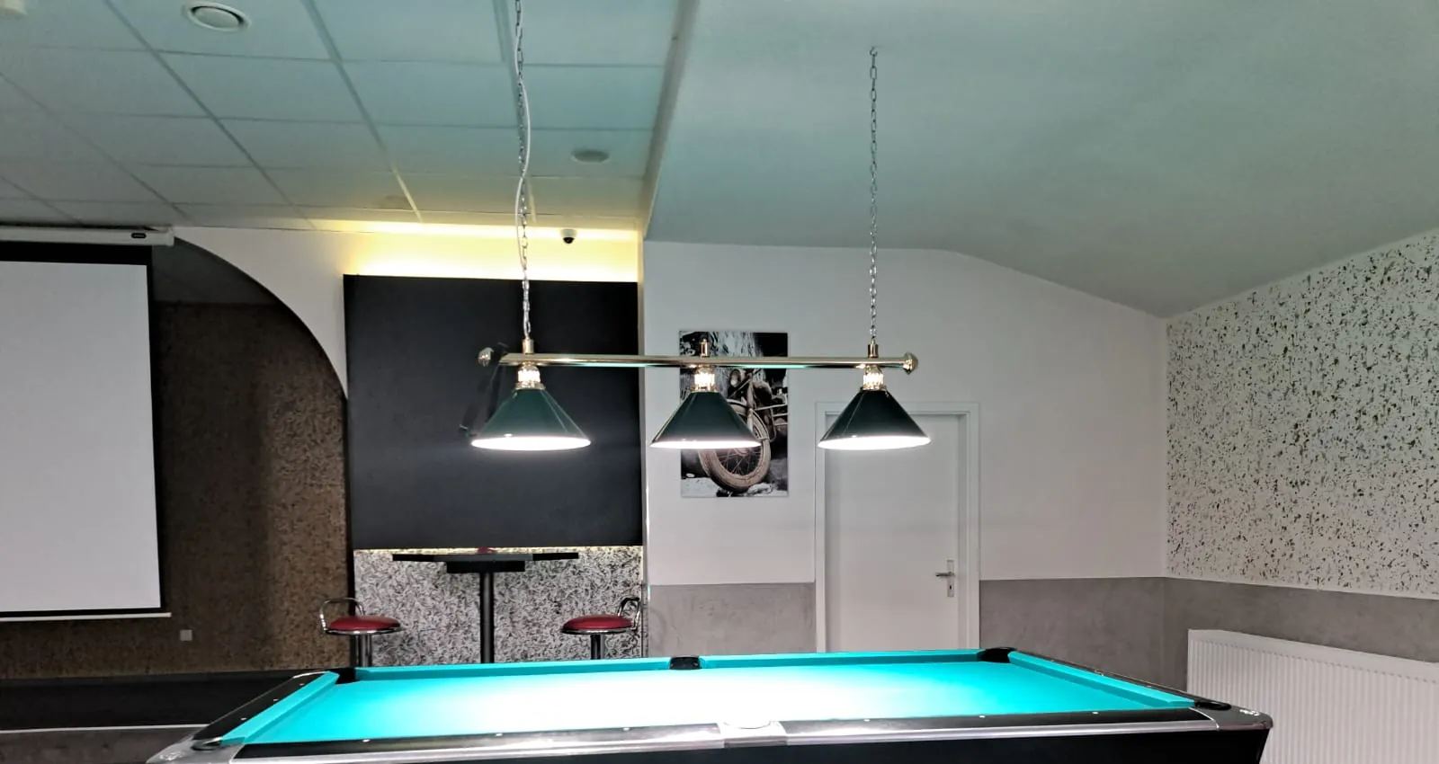 Leinwand an der Wand neben einem Pooltisch mit türkisfarbenem Tuch unter grünen Billardlampen