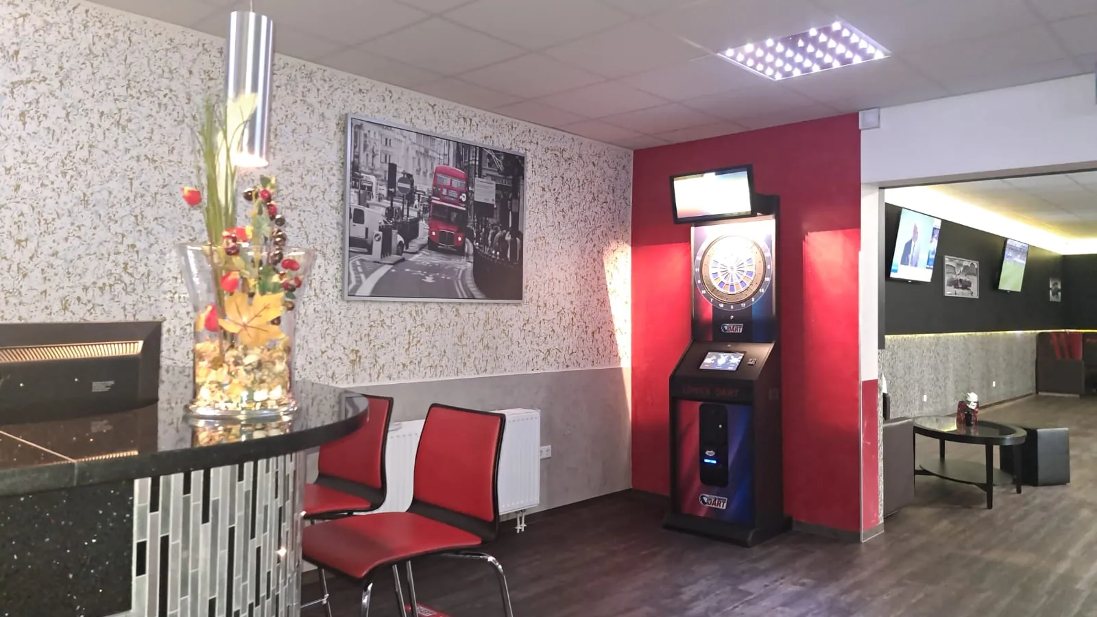 E-Dart-Automat mit Monitor an einer roten Wand der Sportsbar Wesel, davor rote Stühle, links die Ecke der Bartheke