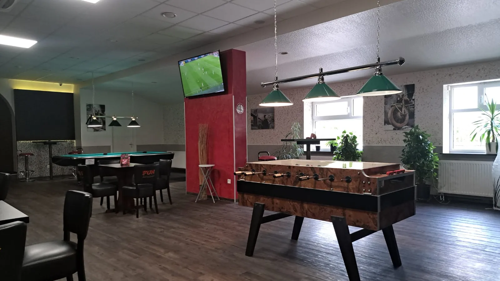 Kicker im Holzdekor vor einer roten Säule mit Fernseher, dahinter Pooltische unter grünen Lampen und Sitzgruppen